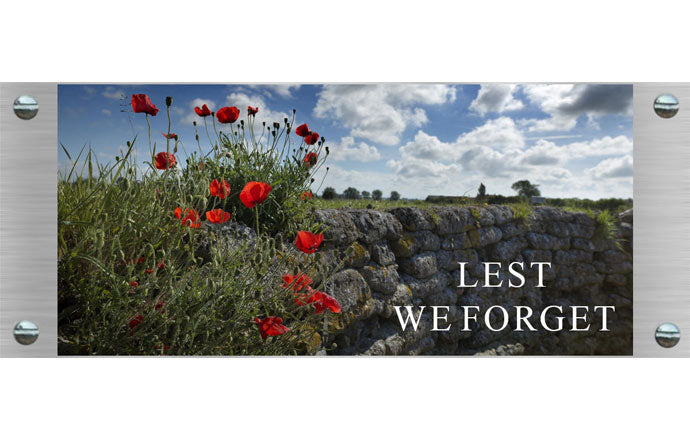 photo memorial with red poppies growing on a stone wall with 'Lest We Forget' text against a blue sky.