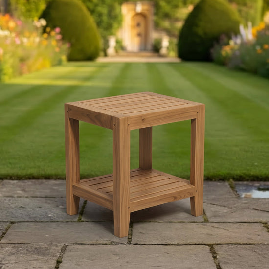 A square wooden teak side table with a slatted top and lower shelf, placed on a stone patio in front of a lush green garden lawn and house.