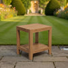 A square wooden teak side table with a slatted top and lower shelf, placed on a stone patio in front of a lush green garden lawn and house.