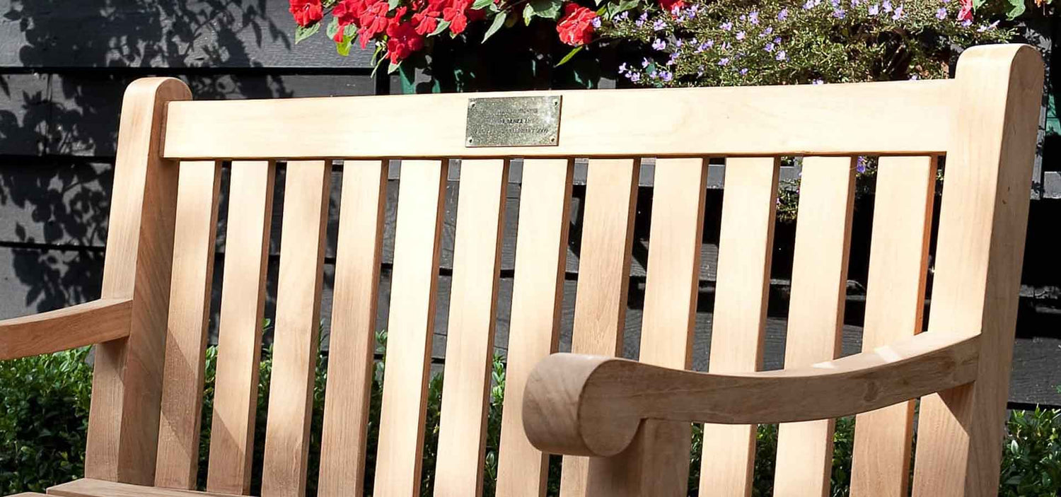 Wooden memorial bench in a garden setting, brass plaque with flowers and a fence in the background
