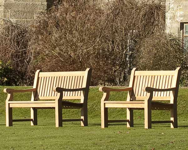 Two traditional wooden memorial benches on a grassy area with a stone building and bushes in the background.